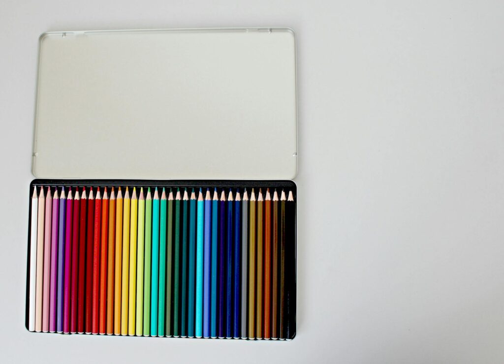 An open tin of coloured pencils arranged in a gradient, set on a pale background with generous blank space.